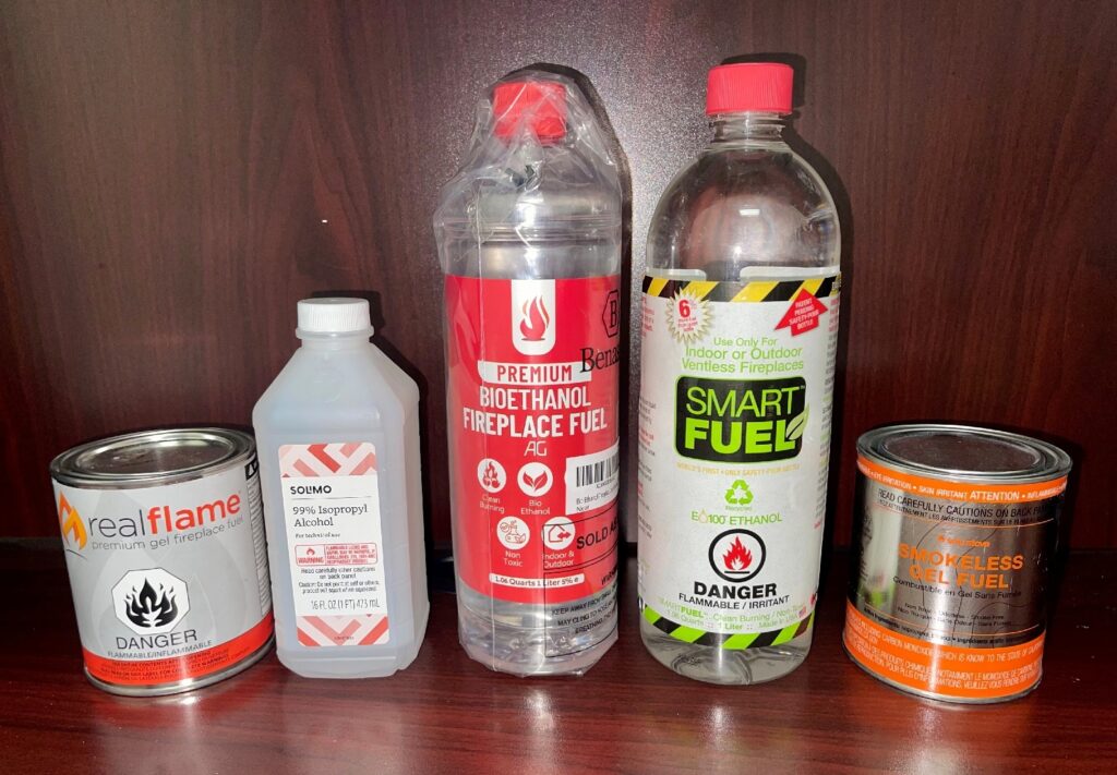 Different alcohol-based fuels shown side by side—rubbing alcohol, ethanol, bioethanol, and gel fuel cans—used in tabletop fire pits